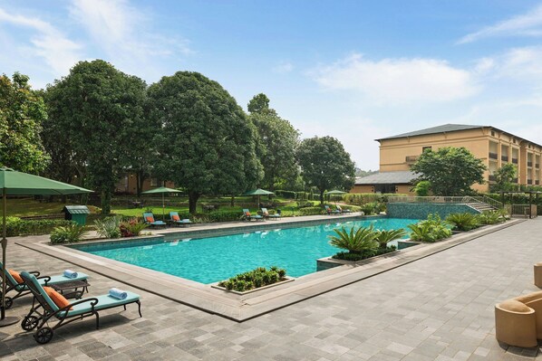 A swimming pool with lounge chairs and umbrellas, surrounded by greenery and a building in the background.