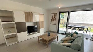 A modern living room with a sofa, TV, and a coffee table. There is a sliding glass door leading to an outdoor area with chairs. - Cocon Neuf 6/8 pers à 2 pas des Plages, Port & Centre Ville (Saint-Pierre-Quiberon)