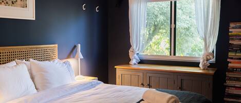 A bedroom with a bed, a nightstand, a lamp, a window with curtains, and a bookshelf.
