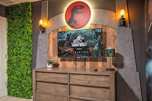 Televisão na parede exibindo 'Jurassic World: Evolution' com logotipo de dinossauro vermelho, um jardim vertical verde e um armário de madeira com gavetas.