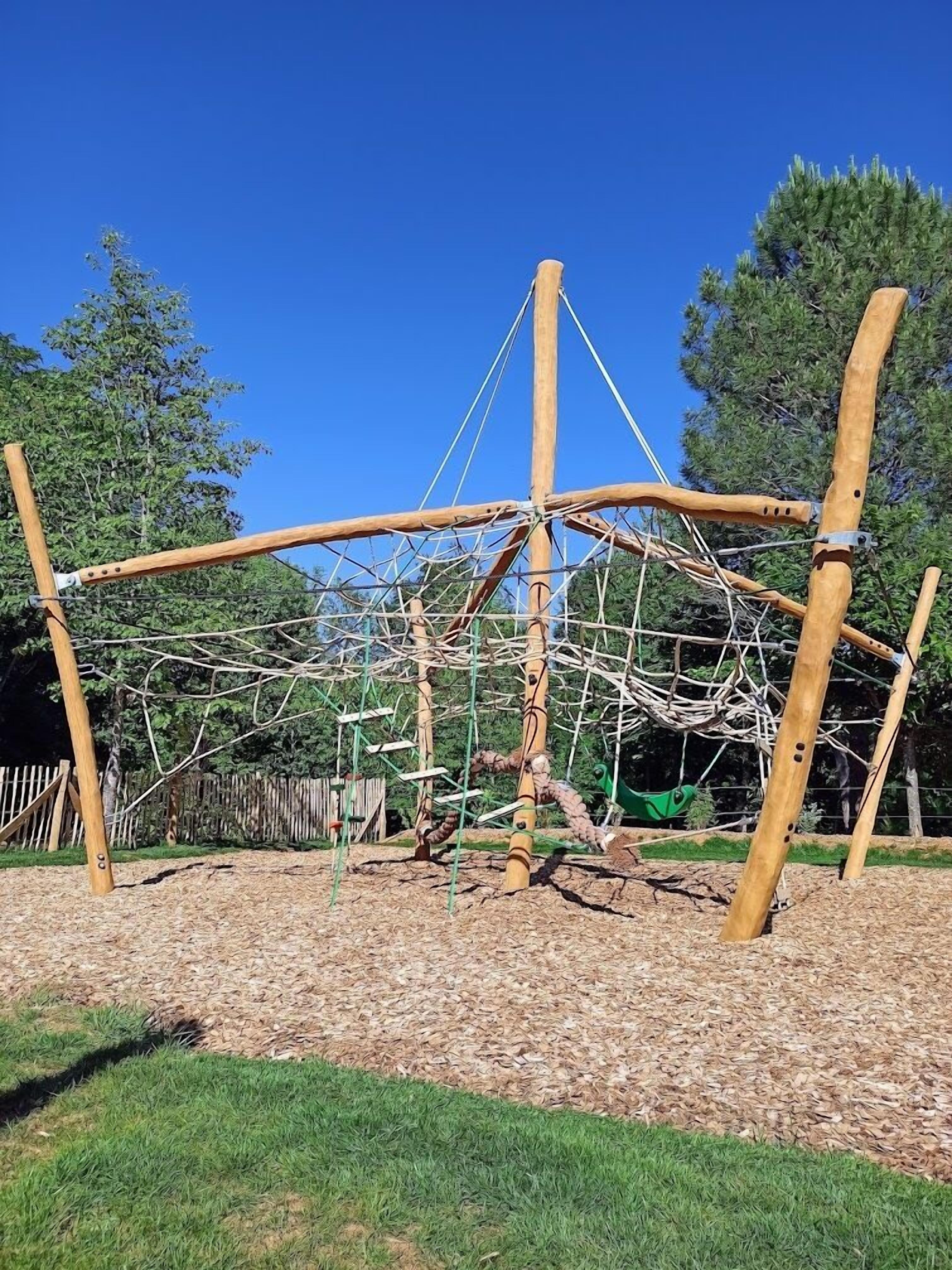 A playground structure made of wooden beams and ropes, surrounded by a grassy area with trees in the background.