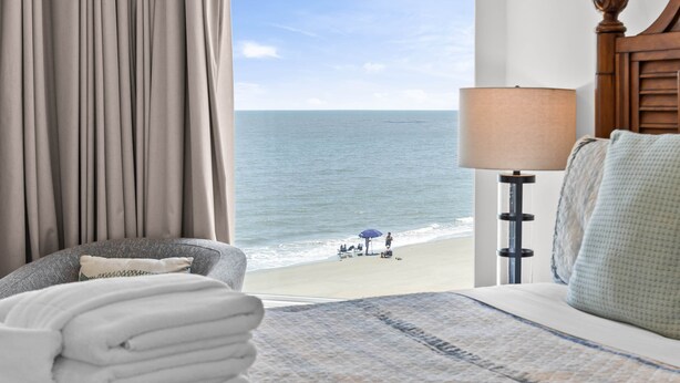 Ein Hotelzimmer mit Bett, Lampe, Blick auf den Strand und einem mit Vorhängen versehenen Fenster.