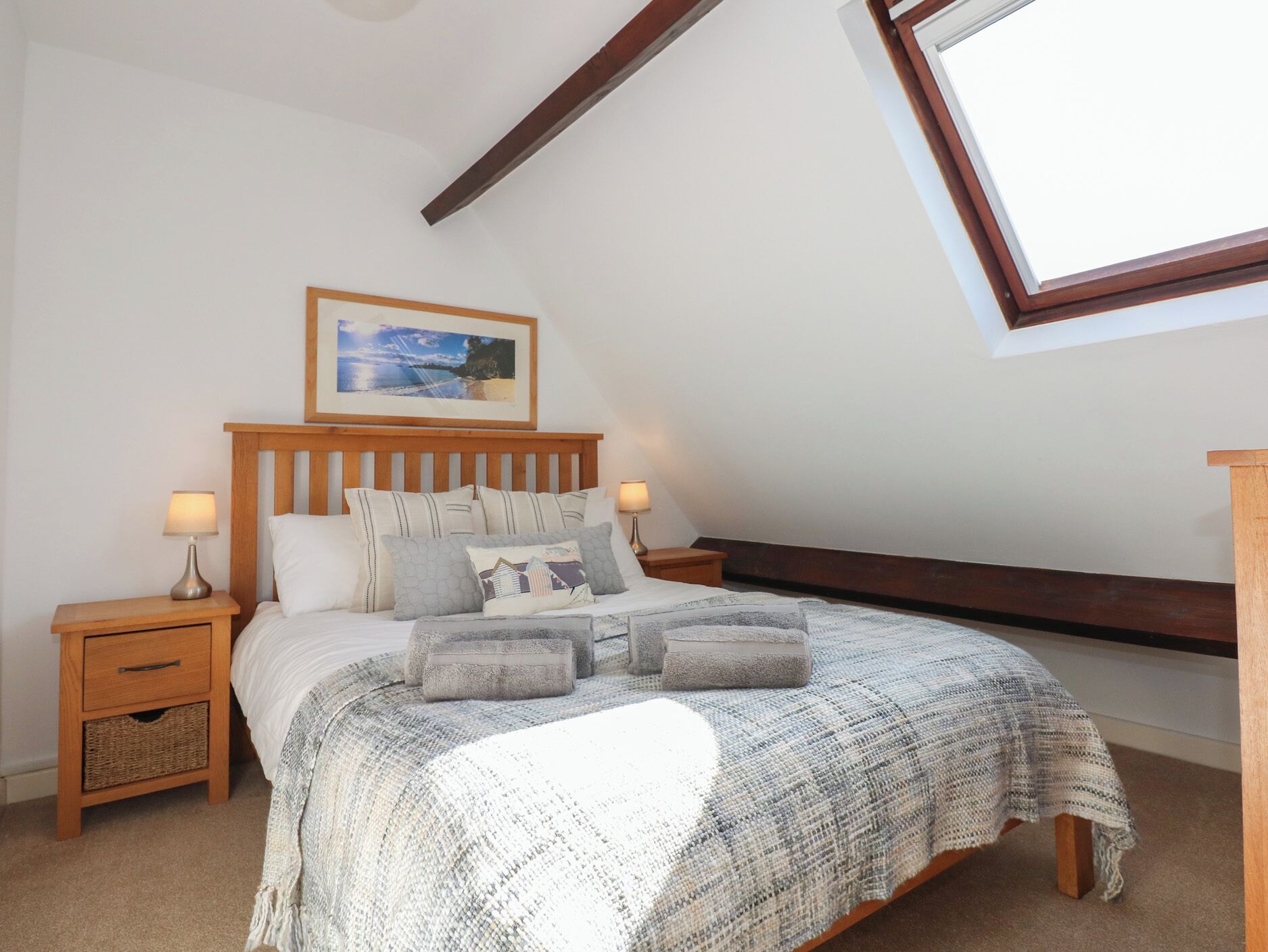 Un dormitorio con techo inclinado, cabecera de madera, cama grande con manta gris de punto, dos mesitas de noche con lámparas y un cuadro enmarcado sobre la cama.