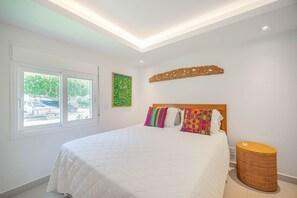 A bedroom with a large bed, a window with a view of the street, and a decorative wall piece. - Bz77 Beachfront house with 4 suites! (Búzios)