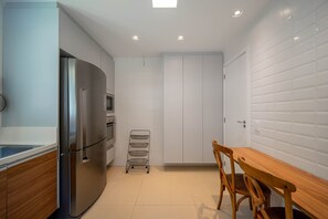 A modern kitchen with a stainless steel refrigerator, wooden cabinets, a small dining table with chairs, and white tiled walls. - Bz77 Beachfront house with 4 suites! (Búzios)