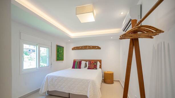 A bedroom with a bed, a standing fan, a window, and a wall-mounted air conditioner. - Bz77 Beachfront house with 4 suites! (Búzios)