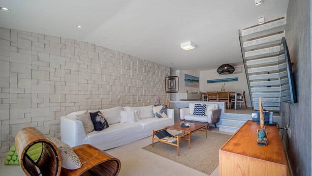 Un salón moderno con un sofá blanco, una mesa de centro de madera y un sillón curvo de madera. Una escalera con barandilla de vidrio conduce a un nivel superior.