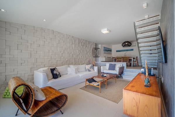 A modern living room with a white sofa, a wooden coffee table, and a curved wooden chair. A staircase with a glass railing leads to an upper level.
