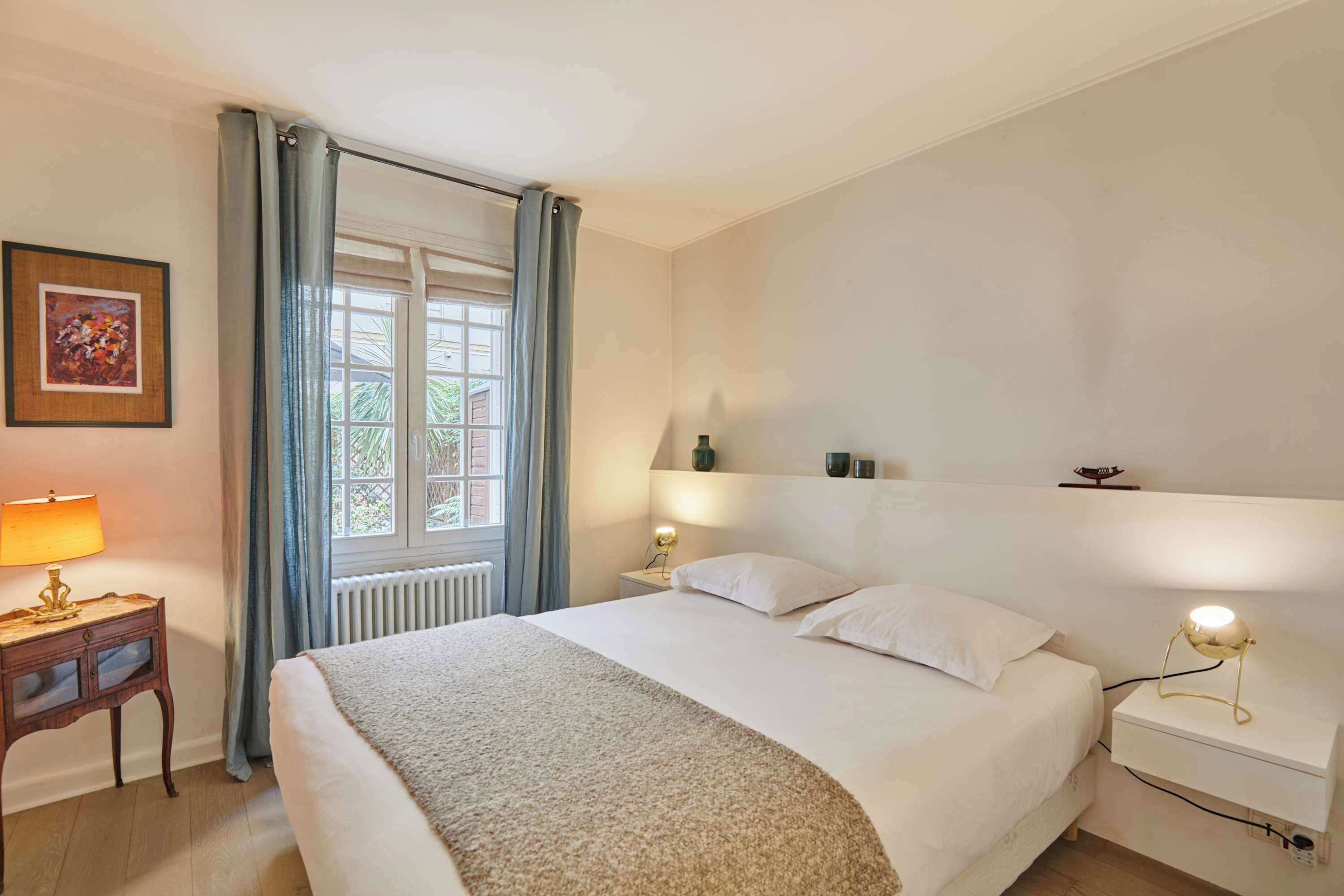 Een slaapkamer met een bed, nachtkastjes, een raam met gordijnen, een wandplank en een lijst met een afbeelding.