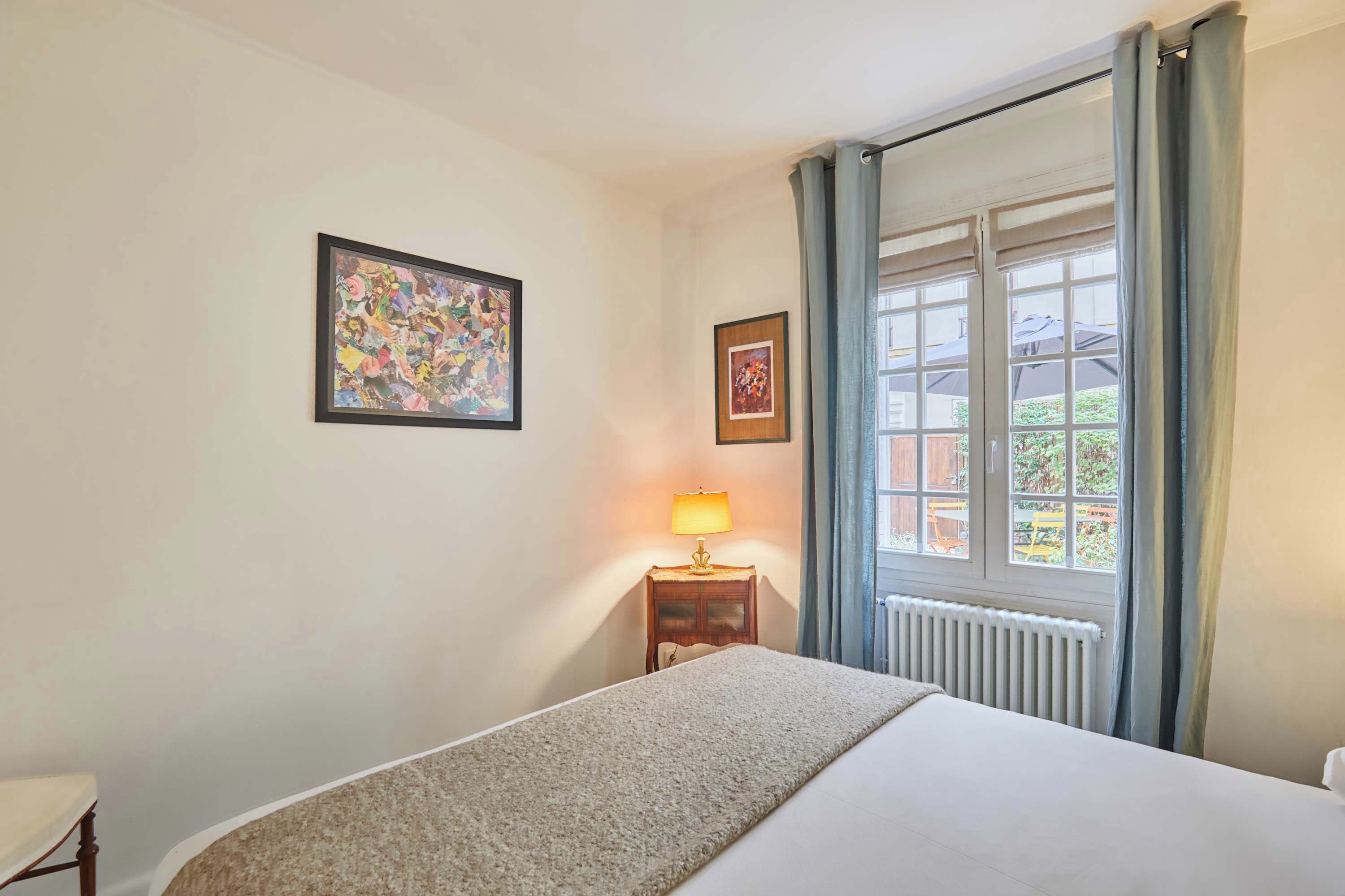 Ein Hotelzimmer mit Bett, Nachttisch mit Lampe, Fenster mit Vorhängen und zwei gerahmten Bildern an der Wand.