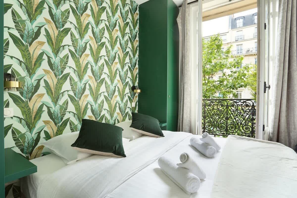 A hotel room with a bed, pillows, a window with a view of buildings, and a green wall with a leaf pattern.
