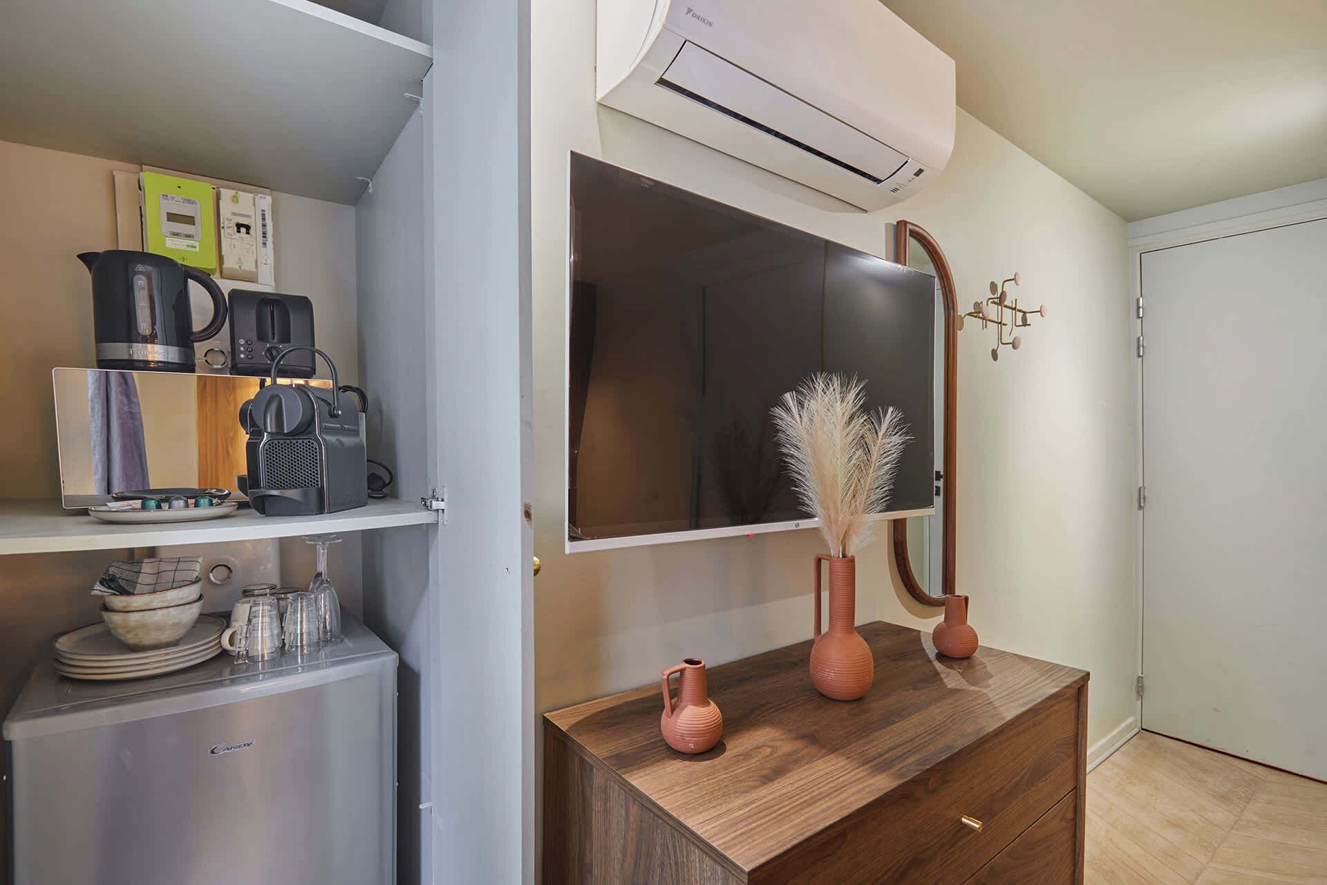 Eine Küchenzeile mit Kaffeemaschine, Mikrowelle und kleinem Kühlschrank. Ein Holzschrank mit zwei Vasen und einem Spiegel. Eine Klimaanlage ist über dem Spiegel angebracht.
