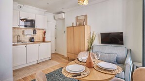 Une cuisine moderne avec des meubles blancs, un four à micro-ondes et une machine à café. On y trouve un coin repas avec une table ronde, des assiettes et un vase rempli de fleurs. On aperçoit également un meuble en bois et un canapé.