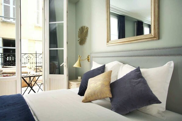 A bedroom with a bed, pillows, a nightstand, a lamp, a mirror, and a view of an outdoor balcony.