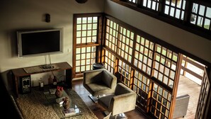 A high-ceilinged living room with a large flat-screen TV, comfortable seating, and a view through a wooden lattice window.