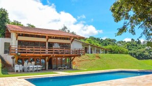 A two-story house with a swimming pool, outdoor seating, and a view of a lush green hillside.