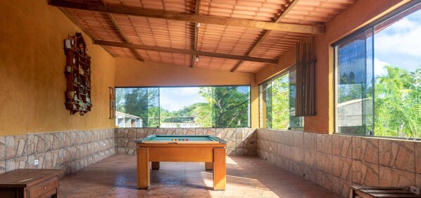 Enjoy a game of pool in this spacious area with a high ceiling and large windows offering views of lush greenery.