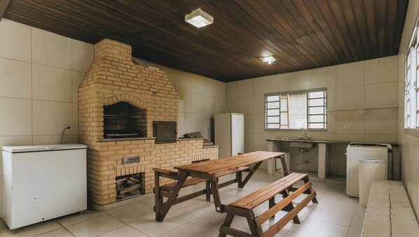 A kitchen with a brick oven, wooden tables, and a window. - Casa em Parelheiros com Piscina e Churrasqueira (São Paulo)
