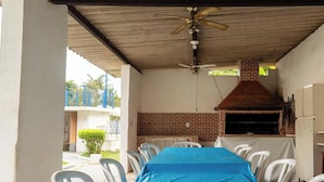 Un patio al aire libre con un horno de ladrillo, sillas de plástico blanco y un mantel azul.