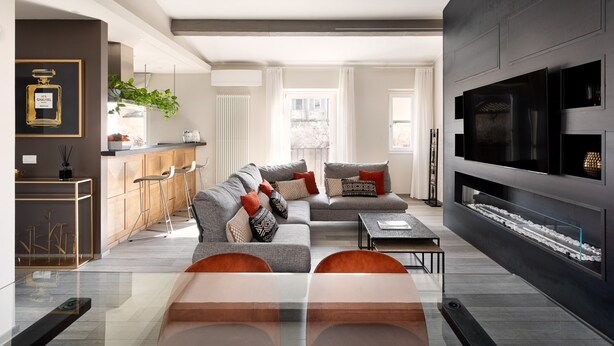 Una sala de estar moderna con sofá, mesa de centro y televisor. Se observa una zona de cocina con taburetes altos y una planta.