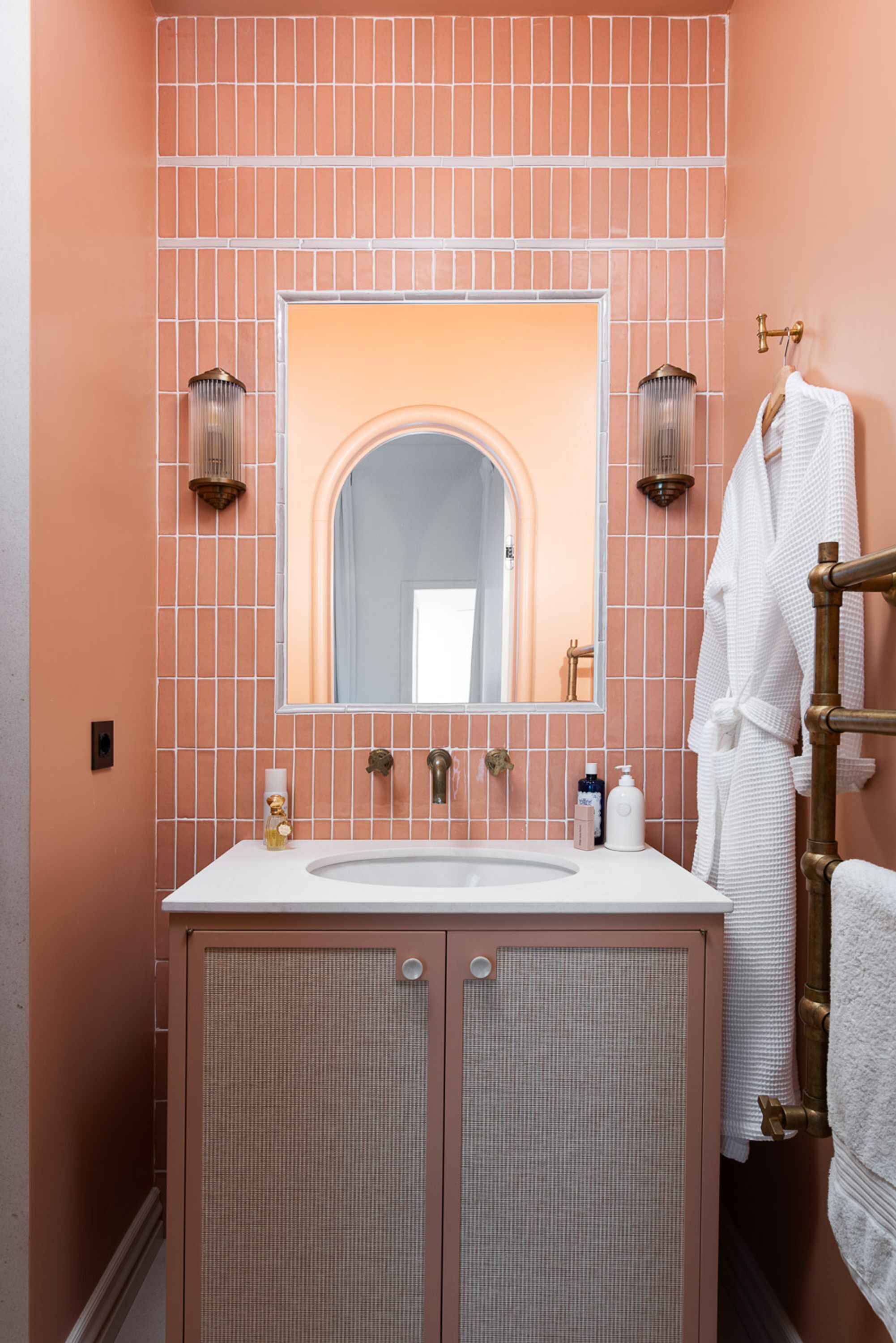 A bathroom with a pink tiled wall, a large mirror, a white sink, two wall-mounted lights, and a towel rack with a white robe.