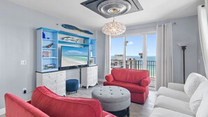 Un salon moderne doté d’un grand téléviseur, de canapés rouges et blancs, d’un pouf gris et d’un balcon offrant une vue sur la mer.