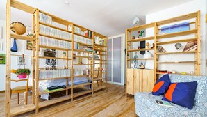 Um quarto com chão de madeira, uma cama com roupa de cama azul e laranja, e duas estantes de madeira repletas de livros e objetos decorativos.