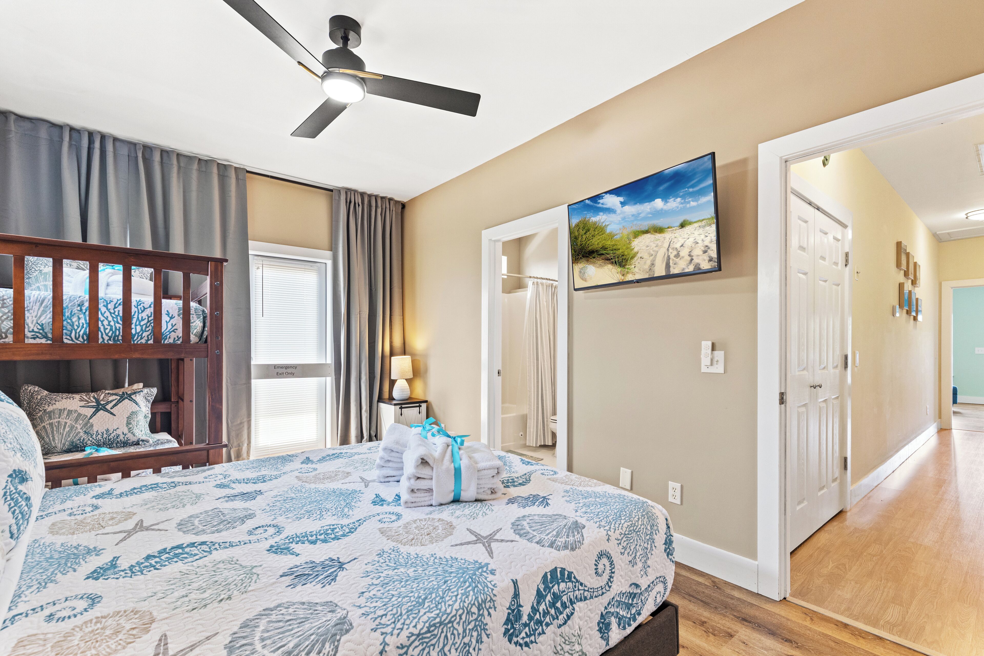 A bedroom with a bed, a ceiling fan, a beach-themed bedspread, a wall-mounted TV, and a door leading to another room.