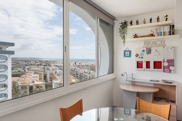 A modern apartment with a large window offering a city view, a small kitchenette, and a dining area with a glass table.