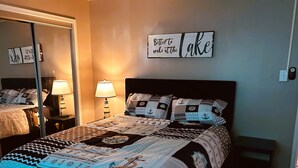 A hotel room with a bed featuring a nautical-themed bedspread, two bedside lamps, and a wall sign that reads "Better to wake at the lake."
