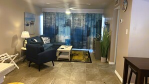 A living room with a sofa, a coffee table, a lamp, and a large window with curtains.