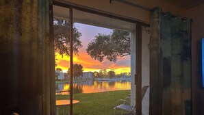 A room with a sliding glass door leading to a patio with a view of a sunset over a lake, a lawn, and a building in the background.