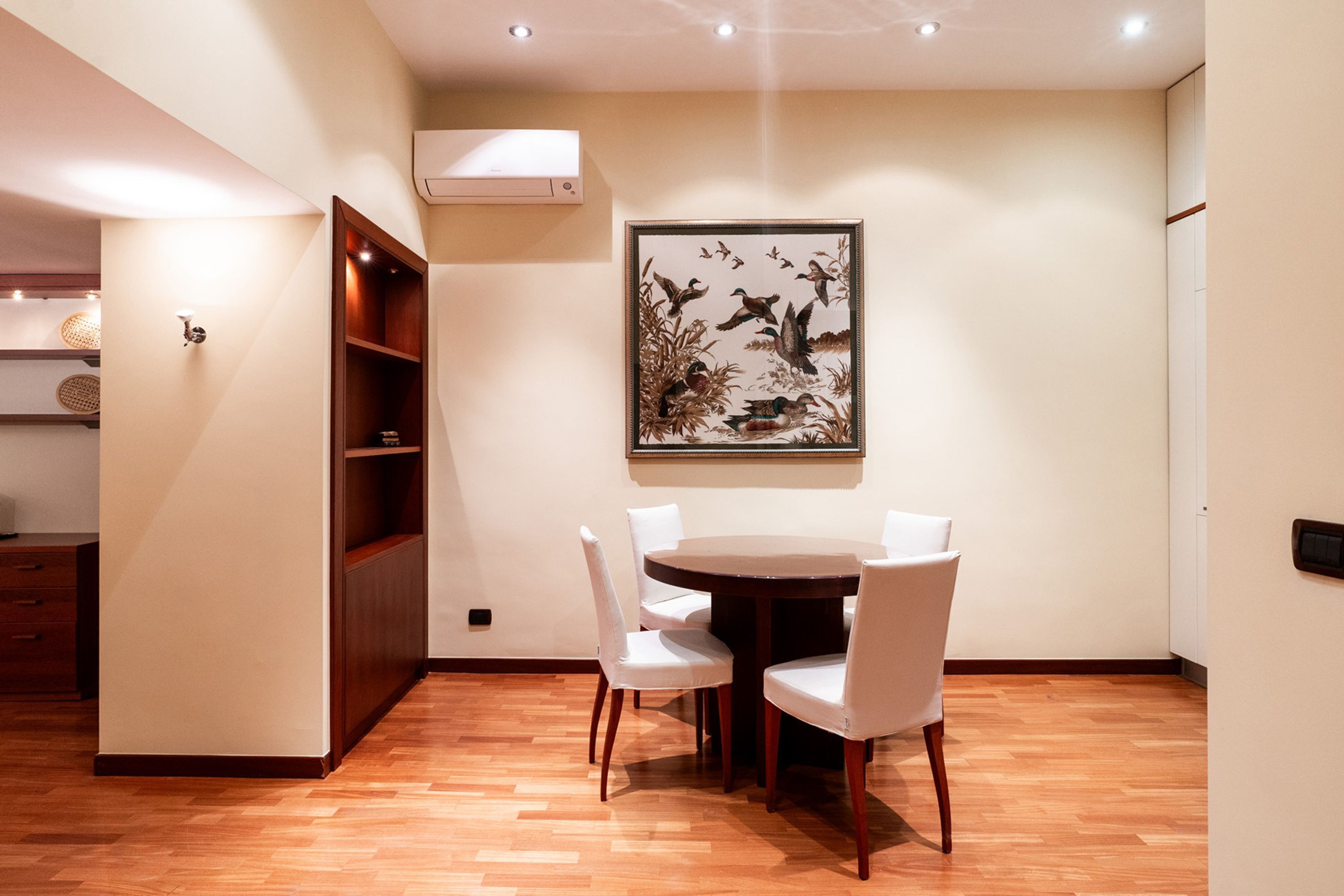 Un comedor con una mesa redonda y cuatro sillas blancas, un armario de madera y una obra de arte enmarcada en la pared.
