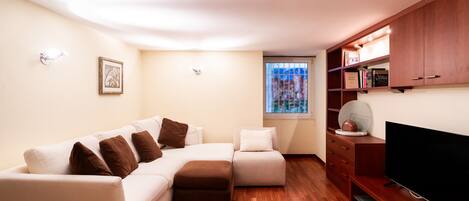 Una sala moderna con un sofá modular, un pequeño reposapiés, un televisor de pantalla plana, piso de madera, una estantería empotrada y una ventana con persianas.