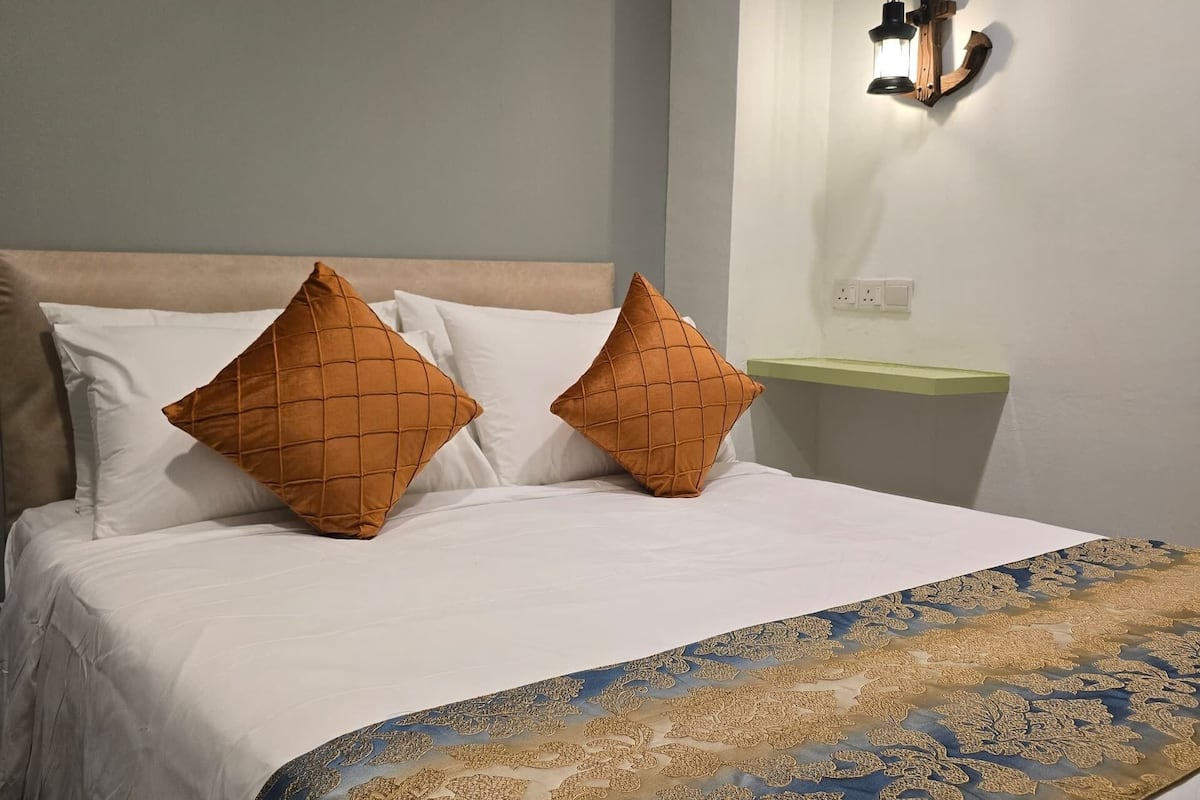 Una cama bien hecha con almohadas blancas y naranjas, una colcha de tonos azules y dorados, y una luz nocturna fijada a la pared.