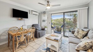 A living room with a TV, ceiling fan, dining table, chairs, sofa, and a balcony with a view. - Beachfront Condo with Communal Pool & Ocean Views! (Hilton Head Island)