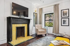 A modern living room with a fireplace, a television mounted above it, a comfortable armchair, and a window with curtains. - Stylish Group Retreat –Sleeps 16 (New York)