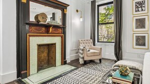 A room with a fireplace, a large window with curtains, a chair, and framed pictures on the wall.