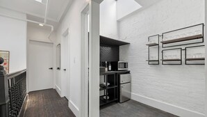A narrow hallway with a dark wooden floor, white walls, and a black metal railing. There is a small kitchenette area with a microwave, a refrigerator, and a shelf with glassware.