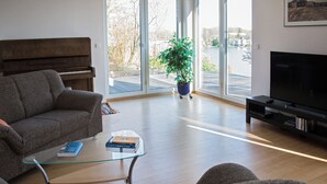 Ein Wohnzimmer mit Klavier, einer Couch, einem Couchtisch mit Büchern, einem Fernsehschrank und einem Blumentopf.