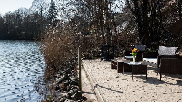 Ein Sitzbereich am Seeufer mit Korbmöbeln und einem Tisch, umgeben von Bäumen und einem Sandweg.