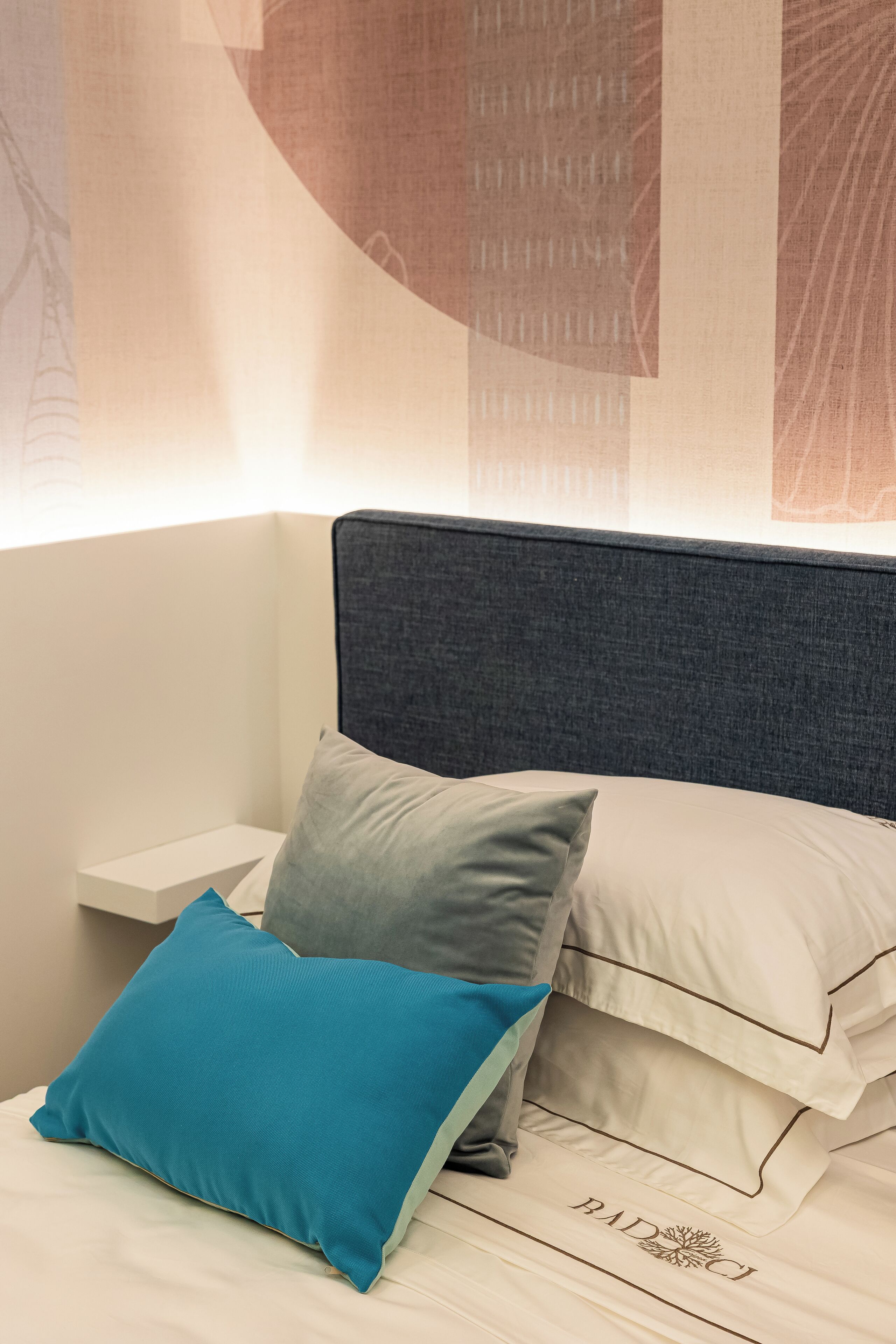 Una cama bien hecha con un cojín azul y uno blanco, un cabecero gris y una mesita de noche blanca.