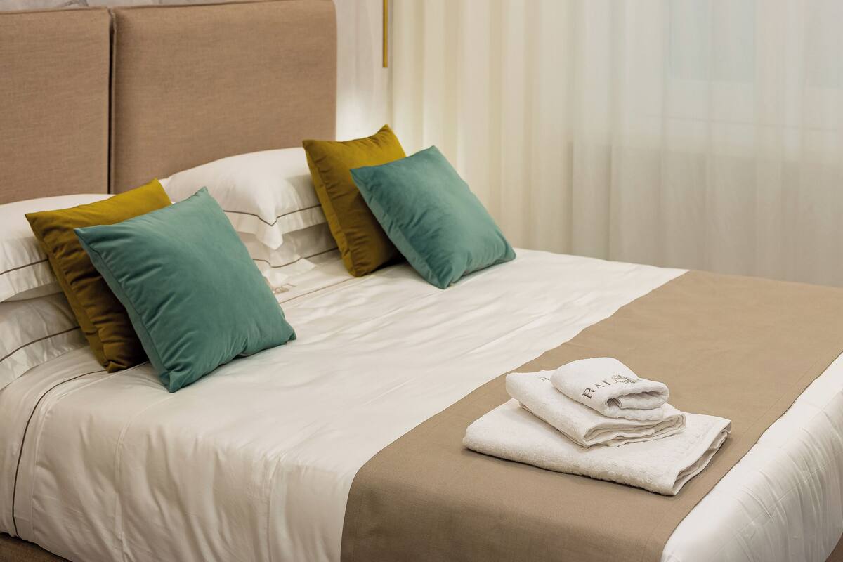 Una cama impecablemente tendida, con un cabecero color beige, ropa de cama blanca y almohadas verde azulado y mostaza.