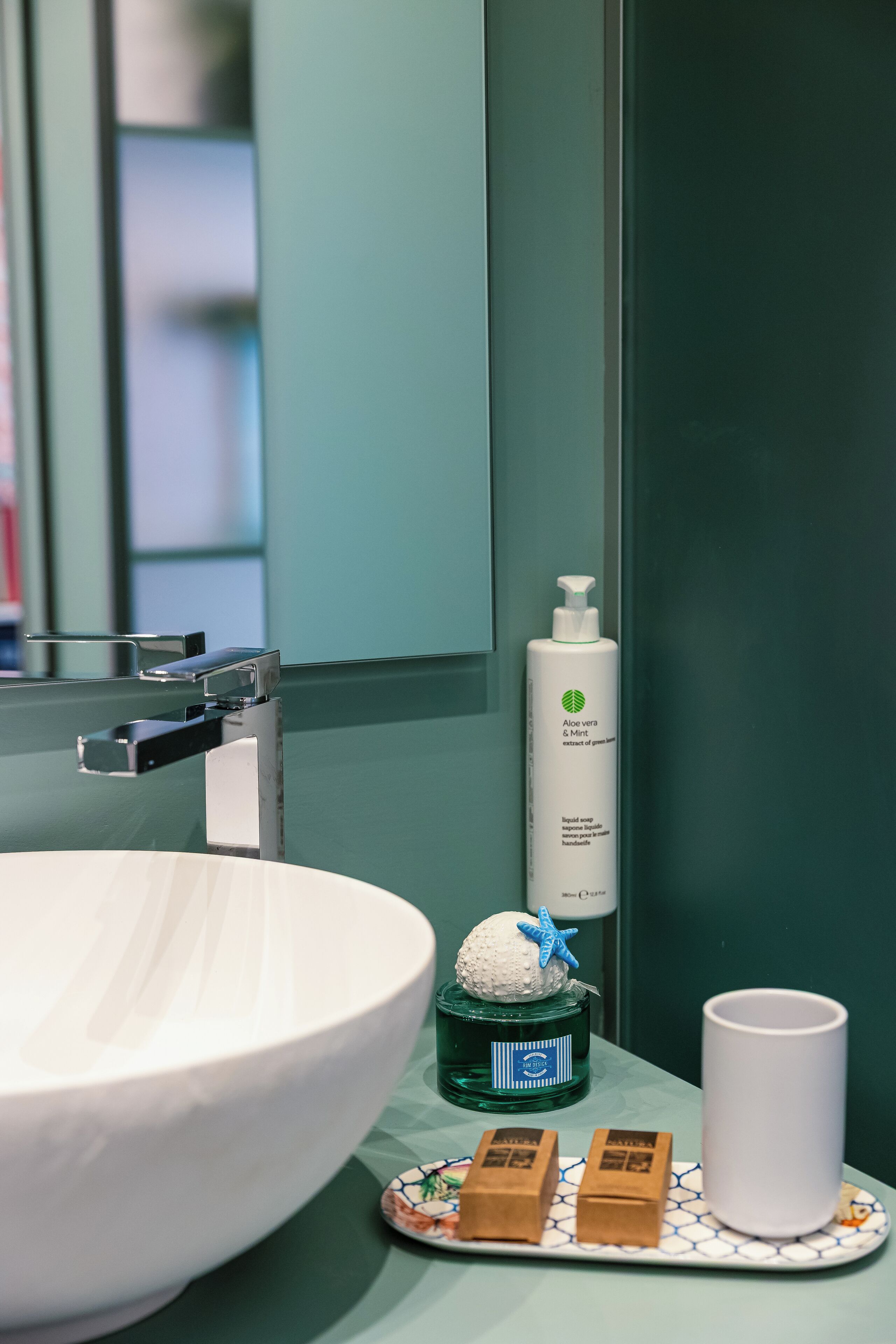 Un baño con un lavamanos blanco, una botella verde de jabón de manos Aloe Vera y Menta, y dos pastillas de jabón marrones sobre un plato decorativo.
