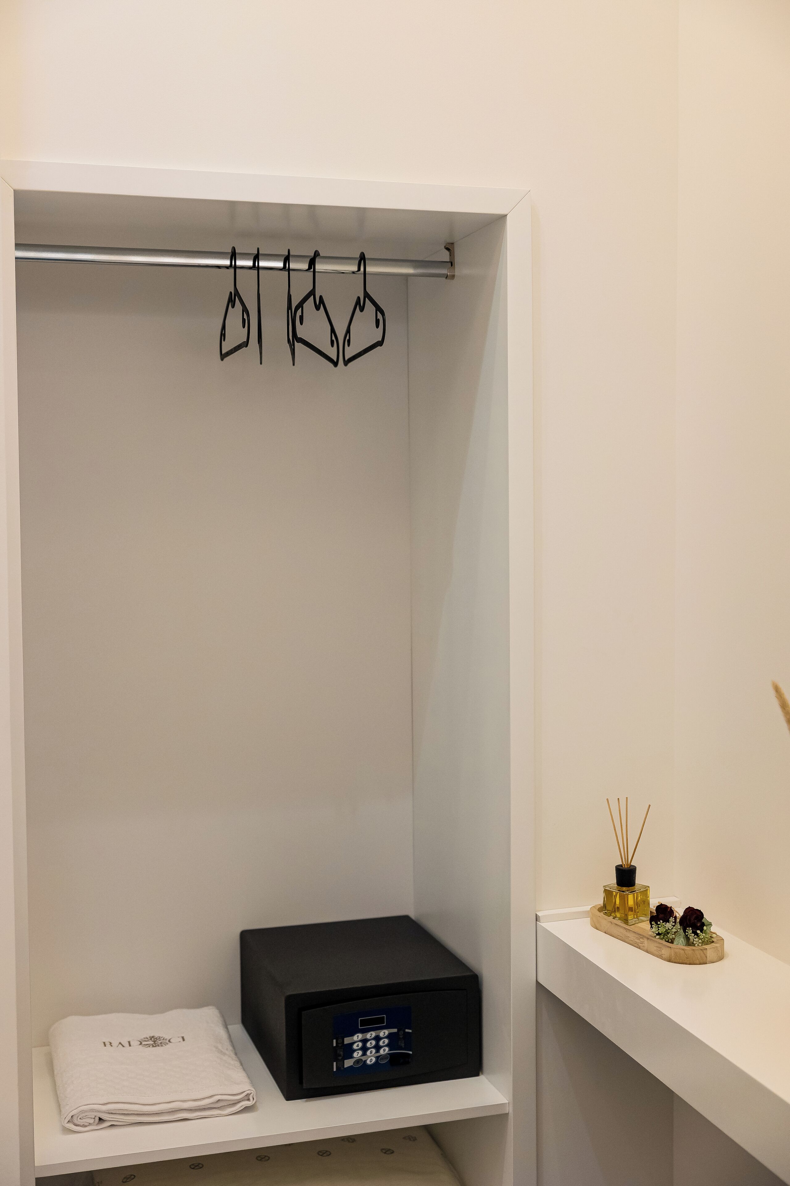 Et pænt organiseret klædeskab med plads til at hænge tøj, et foldet håndklæde, en sort elektronisk enhed og en lille hylde med en diffuser og pynt.