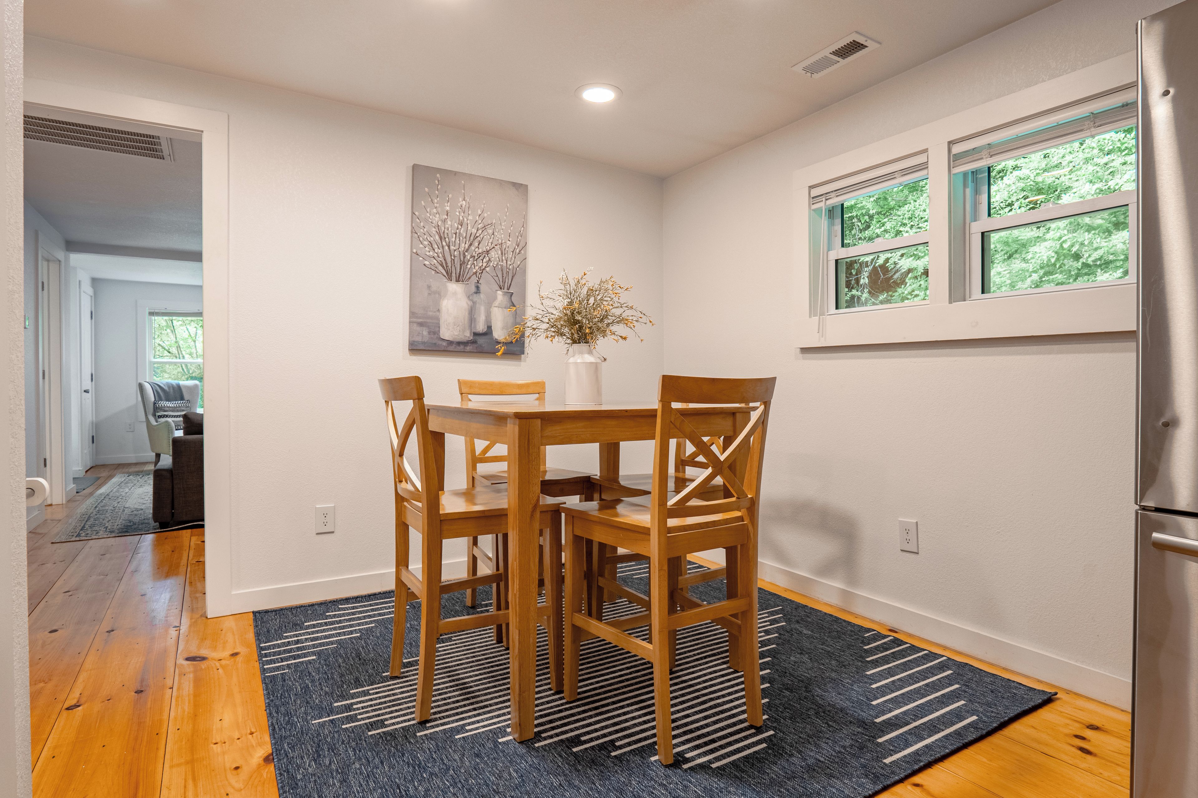 A dining area with a wooden table and chairs, a painting on the wall, and a window with a view of greenery.
