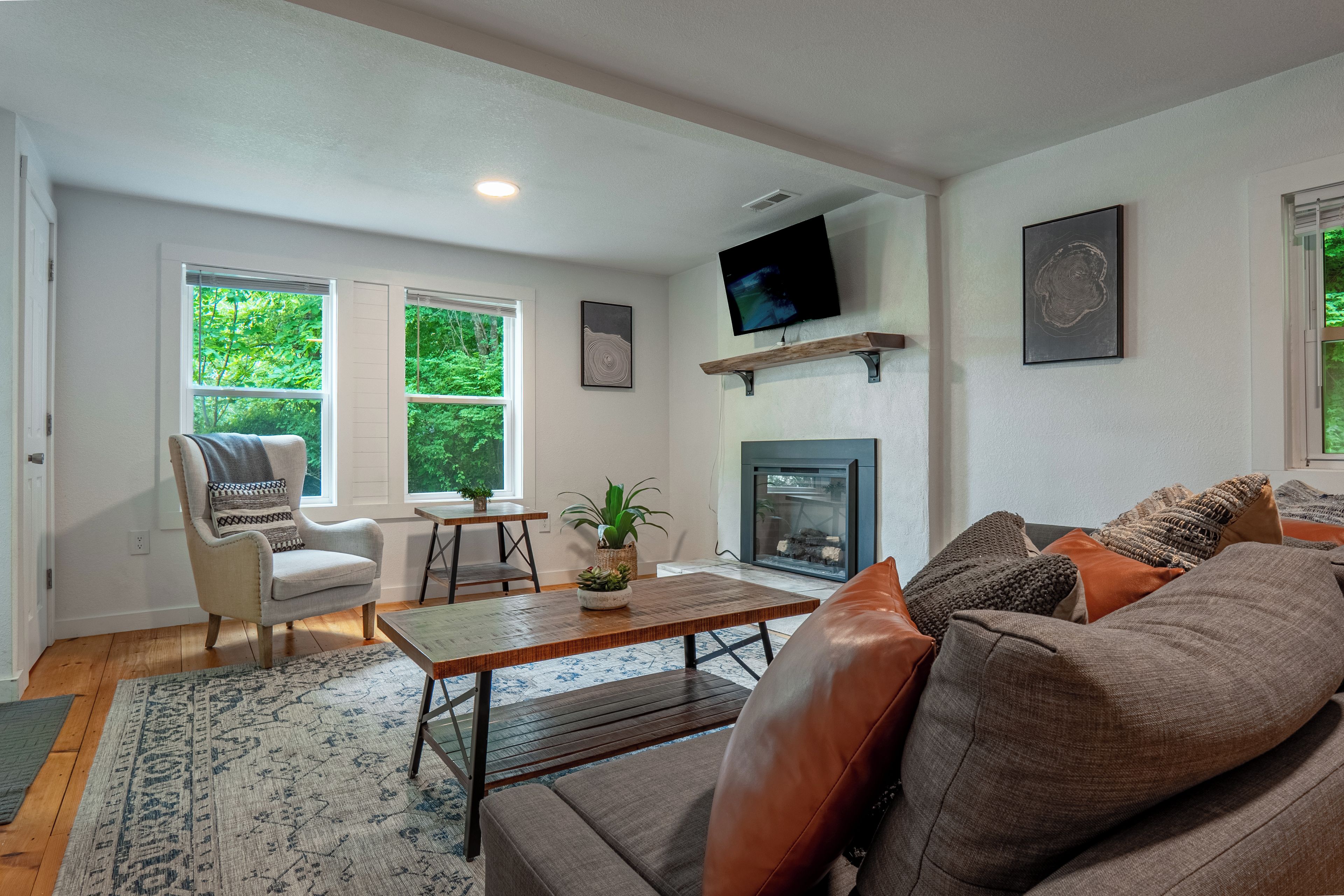 A living room with a fireplace, a television mounted on the wall, and a large window with a view of trees.