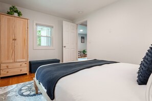 Quarto com cama, guarda-roupa de madeira, janela e porta que dá para outro cômodo.