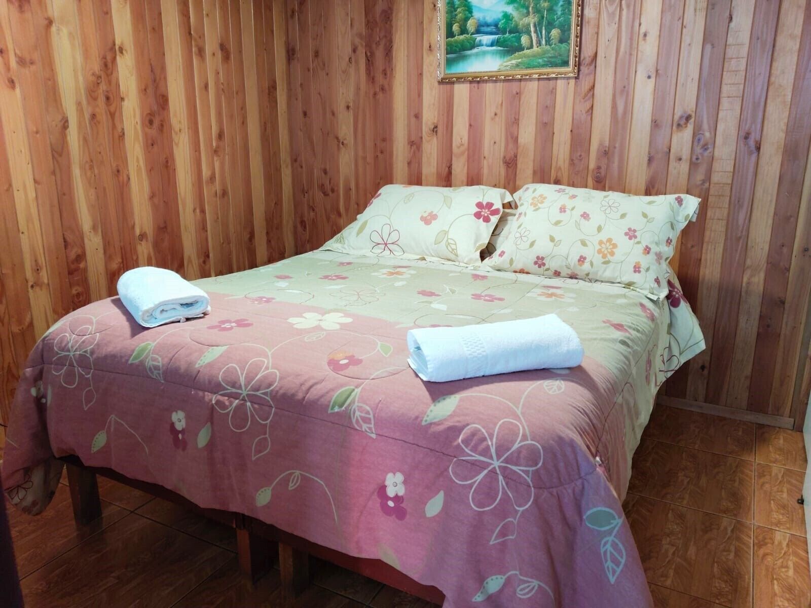 A bed with floral bedding and two pillows, a wooden wall, and a framed picture above the bed.
