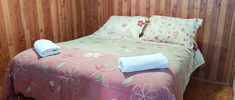 A bed with floral bedding and two pillows, a wooden wall, and a framed picture above the bed.